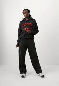 Kobieta stoi w czarnej bluzie z kapturem z napisem "MONTE CARLO" w czerwonych literach, w dopasowanych czarnych spodniach i białych sneakersach, trzymając sznurki od bluzy.