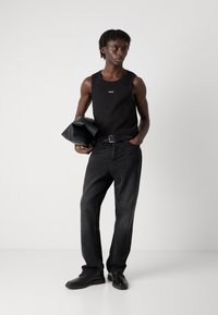 Tricou fără mâneci, negru, cu siglă, asortat cu blugi negri, largi. Ținând o geantă din piele neagră, purtând pantofi negri. Estetică simplă, modernă.