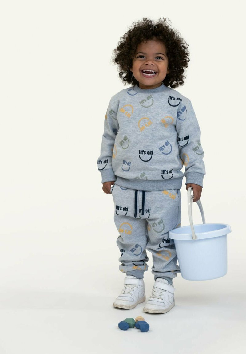 Completo in cotone grigio composto da felpa e pantaloni con design di faccine multicolori, abbinato a sneaker bianche. Bambino che tiene un secchiello di colore azzurro chiaro.