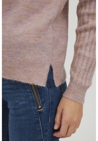 Maglione di un morbido viola chiaro con texture, maniche a coste e spacco laterale. Indossato sopra jeans blu scuri con un evidente dettaglio in zip dorata.