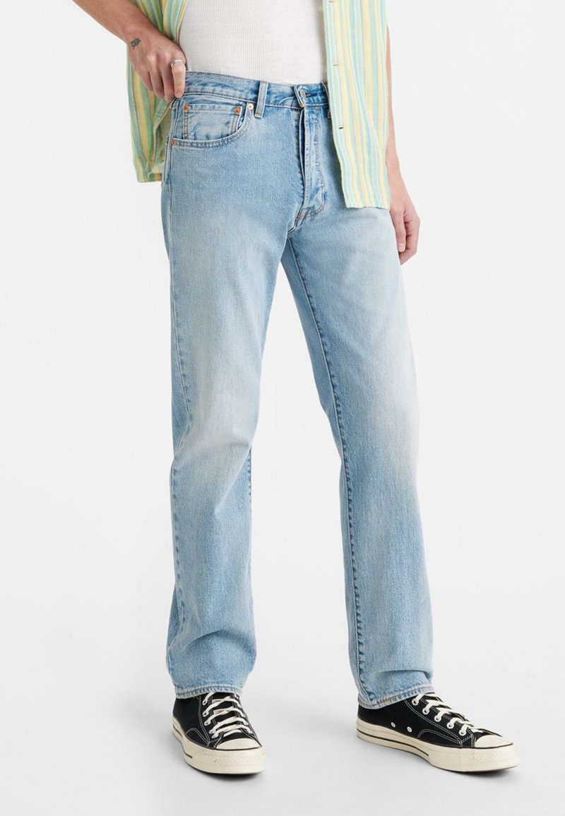 Persona vistiendo jeans rectos de color azul claro, zapatillas negras y blancas, una camiseta blanca y una camisa a rayas verde y amarilla.