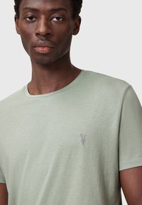Camiseta de algodón verde claro con cuello redondo, que presenta un pequeño diseño bordado de una calavera de chivo en el lado izquierdo del pecho. Textura suave y ajuste casual.
