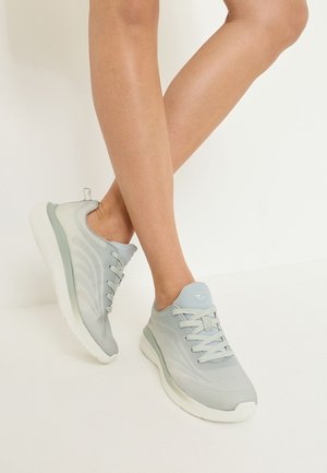 Piernas que llevan zapatillas deportivas gris claro con suelas blancas, de pie sobre un fondo liso color blanco roto.