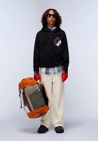 Felpa nera con cappuccio e logo, camicia a scacchi, pantaloni beige larghi, guanti rossi. Porta uno zaino multicolore con dettagli arancioni e testo.