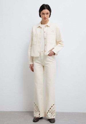 Frau steht vor einer weißen Wand, trägt eine cremefarbene Jacke, dazu passende hochtaillierte Hosen mit ausgeschnittenen Blumenmustern und dunkle Schuhe.