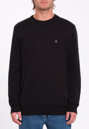 Schwarzer Pullover aus Strickstoff mit Rundhalsausschnitt, gerippten Bündchen und kleinem Logo auf der Brust. Kombiniert mit blauen Jeans.