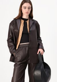 Smeđa kožna overshirt uparen s bež kardiganom i crnom majicom, upotpunjen crnom kožnom torbom i odgovarajućim hlačama.