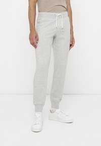 Homme portant un pantalon de survêtement gris clair avec taille à cordon et des baskets blanches, debout devant un fond blanc uni.