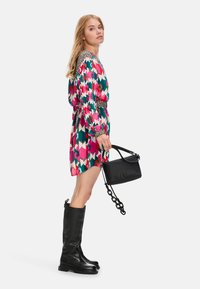 Bunt gefärbtes Kleid mit einem geometrischen Muster in Pink, Grün und Creme, ergänzt durch einen schwarzen Gürtel, kombiniert mit hohen schwarzen Stiefeln und einer schwarzen Handtasche.