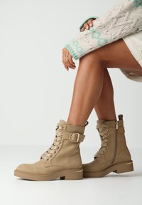 Beige kožené kotníkové boty s šněrováním vpředu, bočním zipem, dekorační přezkou a blokovým podpatkem se zlatým akcentem. Texturovaný povrch.