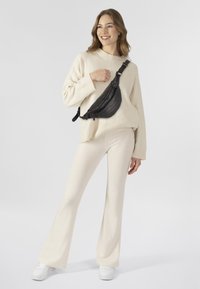 Pullover aus cremefarbenem Strick, ausgestellte cremefarbene Hose und schwarze Leder-Crossbody-Tasche mit Reißverschluss und Beschlägen. Weiße Sneakers.