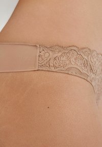 String beige en dentelle avec une ceinture lisse, présentant des motifs en cœur complexes le long de la bordure en dentelle et une finition en tissu doux et sans couture.