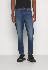 Vaqueros de mezclilla azul oscuro con corte slim, diseño de cinco bolsillos, sutil desvanecimiento y costuras en contraste, llevados con zapatillas negras.