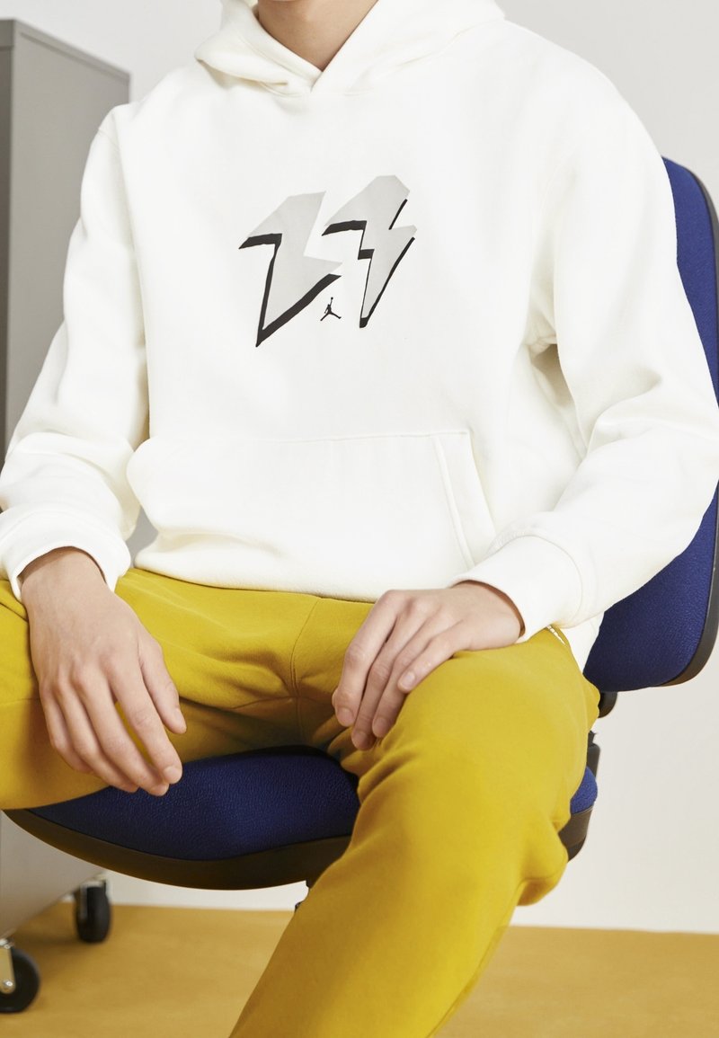 Felpa bianca con cappuccio e design di fulmine nero e grigio sul petto, abbinata a pantaloni giallo senape.