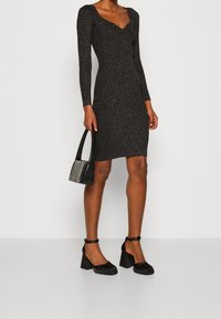 Abito bodycon nero lucido con maniche lunghe e scollatura a V profonda. Abbinato a scarpe nere con tacco e una borsa nera dalla texture.