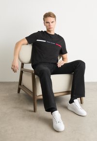 Schwarzes kurzärmliges T-Shirt mit weißen und roten horizontalen Streifen sowie dem "TOMMY HILFIGER"-Logo, kombiniert mit schwarzen Jeans und weißen Sneakers.