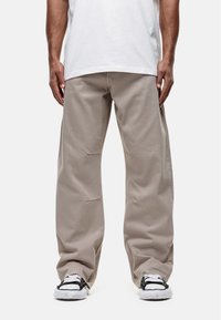 Persoon in losse beige broek en zwart-witte sneakers, staand tegen een eenvoudige witte achtergrond.