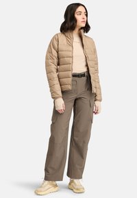 Beige pufferjack met een horizontaal gestikt patroon, gecombineerd met een lichtbruine top met lange mouwen en cargobroek. Crème-kleurige schoenen.