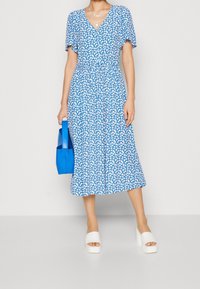 Donna che indossa un vestito midi floreale blu con sandali con la piattaforma bianchi, mentre tiene una borsa blu brillante su uno sfondo neutro.