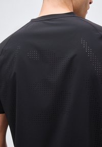 Άντρας που φοράει μαύρο κοντομάνικο πουκάμισο με μικρές διατρήσεις που σχηματίζουν αφηρημένα σχέδια στο πάνω μέρος της πλάτης, σε απλό φόντο.
