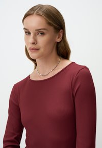 Geribbeld, langwerpig bordeauxrood top met een brede halslijn. Kenmerkt zich door een minimalistisch ontwerp, een gladde textuur en is gecombineerd met een eenvoudige zilveren ketting.