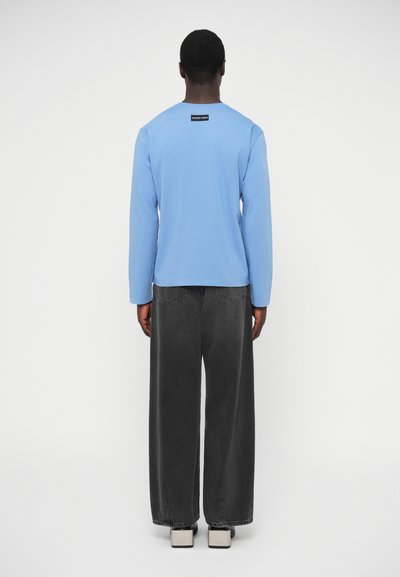 Svetlomodrá košeľa s dlhým rukávom a okrúhlym výstrihom, v kombinácii s širokými tmavosivými džínsami a svetlými topánkami, zobrazená zozadu.