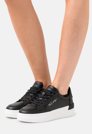 Baskets en cuir noir avec semelles en caoutchouc blanches, dotées de lacets plats, d'un détail logo sur le côté et d'une texture lisse.