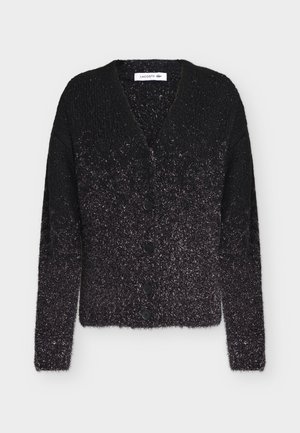 Femme portant un cardigan dégradé noir et gris, un haut blanc, un pantalon large noir, des chaussures noires, tenant un petit sac à main beige.