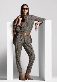 Schwarzer und beigefarbener gemusterter Overall mit Button-up-Kragen, tailliertem Gürtel und braunen Stiefeletten. Accessoires mit Sonnenbrille und Schmuck.