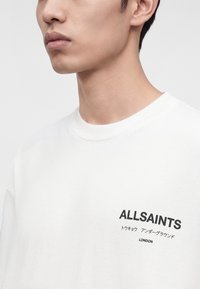 Gros plan d'une personne portant une chemise blanche avec "ALLSAINTS," texte japonais et "LONDON" imprimé sur la poitrine.