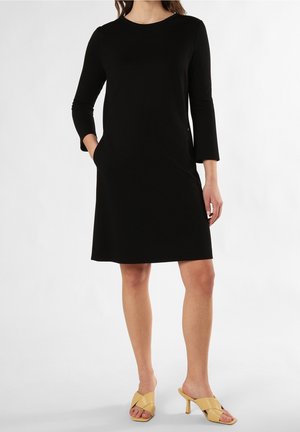 Kobieta w czarnej sukience do kolan z rękawami do łokci i beżowych sandałach na obcasie, pozująca z jedną ręką w kieszeni.