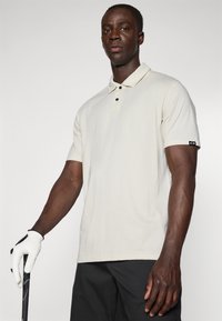 Polo shirt crème à manches courtes avec un col, patte de boutonnage à deux boutons et bord noir sur les manches, assorti à un short de golf noir et des gants.