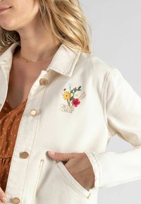 Cremfarbene Denimjacke mit goldfarbenen Knöpfen, die florale Stickereien in Farben wie Gelb, Rot und Blau aufweist. Mit seitlichen Taschen und sichtbaren Nähten.