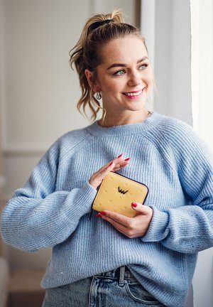 Yellow wallet with textured polka dot design and dark zipper, held against a light blue sweater. Features a logo embossed on the front.
