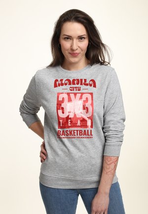 Kobieta z długimi brązowymi włosami nosząca szarą bluzę z czerwonym napisem „Manila City 3x3 Team Basketball Championship Tournament.”
