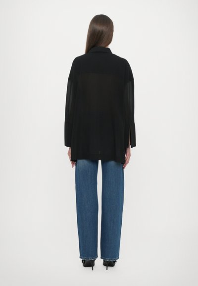 Zwarte doorschijnende blouse met lange mouwen, zijsplitten en een kraag, gecombineerd met recht gesneden blauwe denim jeans en zwarte hoge hakken.