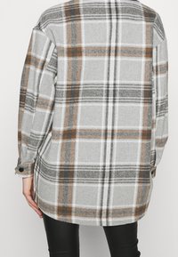 Camisa de gran tamaño con estampado de cuadros gris que presenta líneas blancas y marrones, puños abotonados y textura de tela suave con un diseño de dobladillo curvado.