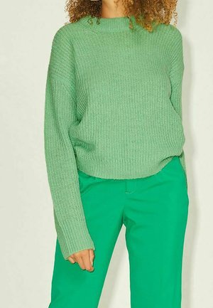 Osoba v světle zeleném pleteném svetru s dlouhými rukávy a jasně zelených kalhotách stojící před neutrálním pozadím.