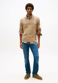 Joven de pie, vistiendo un suéter beige de punto trenzado sobre una camisa de cuadros rojos, jeans azules y zapatillas marrones sobre un fondo blanco.