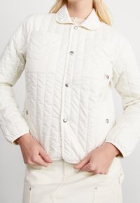 Pehmeä, sileäpintainen vaaleanvärinen tikattu takki, jossa on napillinen edusta, litteä kaulus, kaksi etutaskua ja hopeanväriset koristeosat.