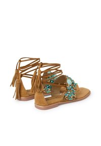 Sandalo in suede marrone con accenti di gemme turchesi, dettagli in frange e suola piatta. Design aperto con fasce incrociate sul piede.