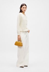 Crème wol cardigan met knopen, witte hooggerende broek, en een kleine gele handtas. De schoenen zijn puntig met een zwarte punt.