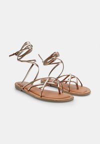 Sandale strappy din bronz metallic cu talpă plată. Bretelele sunt elegante și se învârt în jurul gleznei, contrastând cu talpa de culoare arămie.