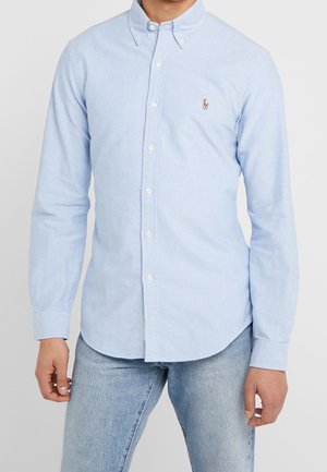 Homme portant une chemise Oxford bleu clair boutonnée avec des manches longues et un petit logo brodé sur la poitrine, associée à un jean bleu clair.