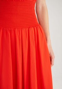 Robe rouge avec un corsage smocké et une jupe fluide, réalisée en tissu léger avec une texture lisse et un design plissé.