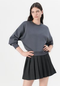 Sweatshirt cinza oversized com mangas longas e punhos canelados, combinado com uma saia preta plissada. Os tecidos são suaves e texturizados.