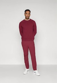 Sudadera deportiva burdeos con cuello acanalado, combinada con pantalones rectos a juego. La modelo lleva zapatillas blancas y está de pie frente a un fondo claro.