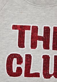 Pilkas audinys, džemperis su „THE CLUB“ raudonais blizgučiais, apžiuožtas baltu siūlu. Apvalus iškirpimas, lygus paviršius, laisvas kirpimas.