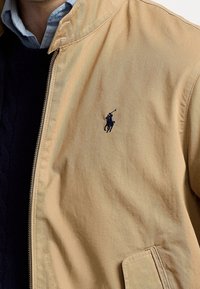 Lichtgewicht beige zip-up jack met subtiel marineblauw logo, gestructureerde kraag en detail met zijzak.