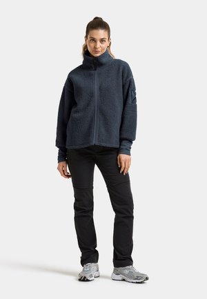 Kvinde stående iført en mørkeblå fleecejakke, sorte bukser og grå sneakers mod en ensfarvet lysegrå baggrund.